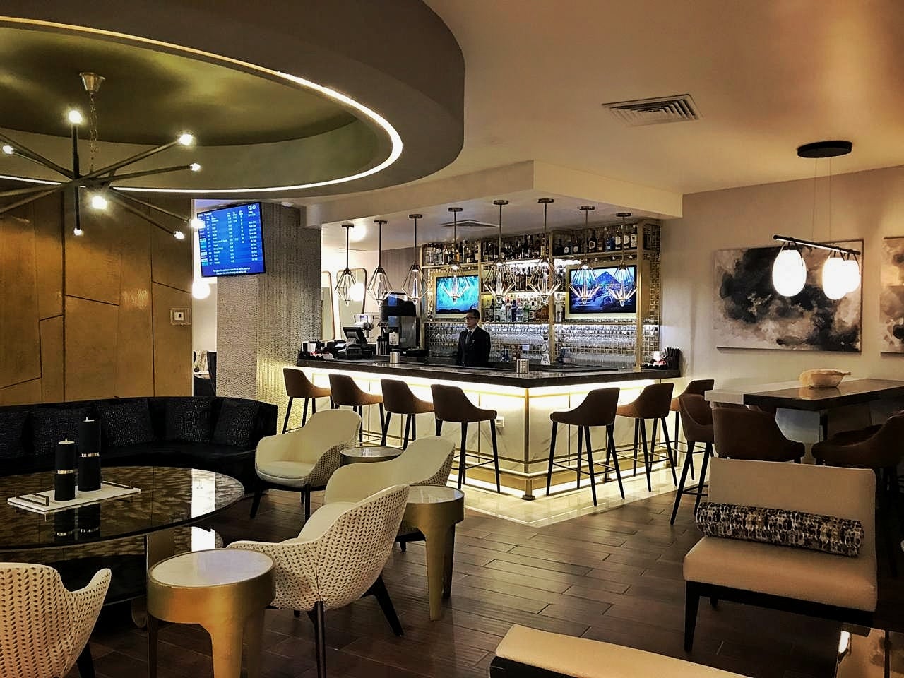 A bar area in the Mera Business Lounge Cancun with a backlit counter, high stools, and a fully stocked display of bottles and glassware behind. The space includes pendant lighting, lounge seating with low tables in the foreground, and a mounted flight information screen on the wall. A bar area in the Mera Business Lounge Cancun with a backlit counter, high stools, and a fully stocked display of bottles and glassware behind. The space includes pendant lighting, lounge seating with low tables in the foreground, and a mounted flight information screen on the wall.