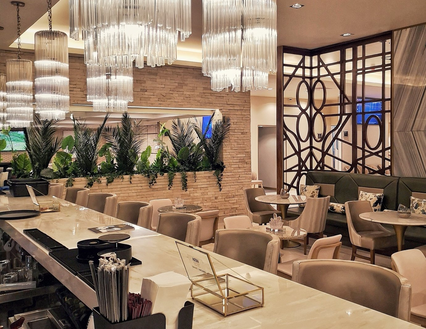 A bar and seating area in the Mera Business Lounge Cancun with a long marble counter, beige upholstered chairs, and small round tables. A bar and seating area in the Mera Business Lounge Cancun with a long marble counter, beige upholstered chairs, and small round tables.