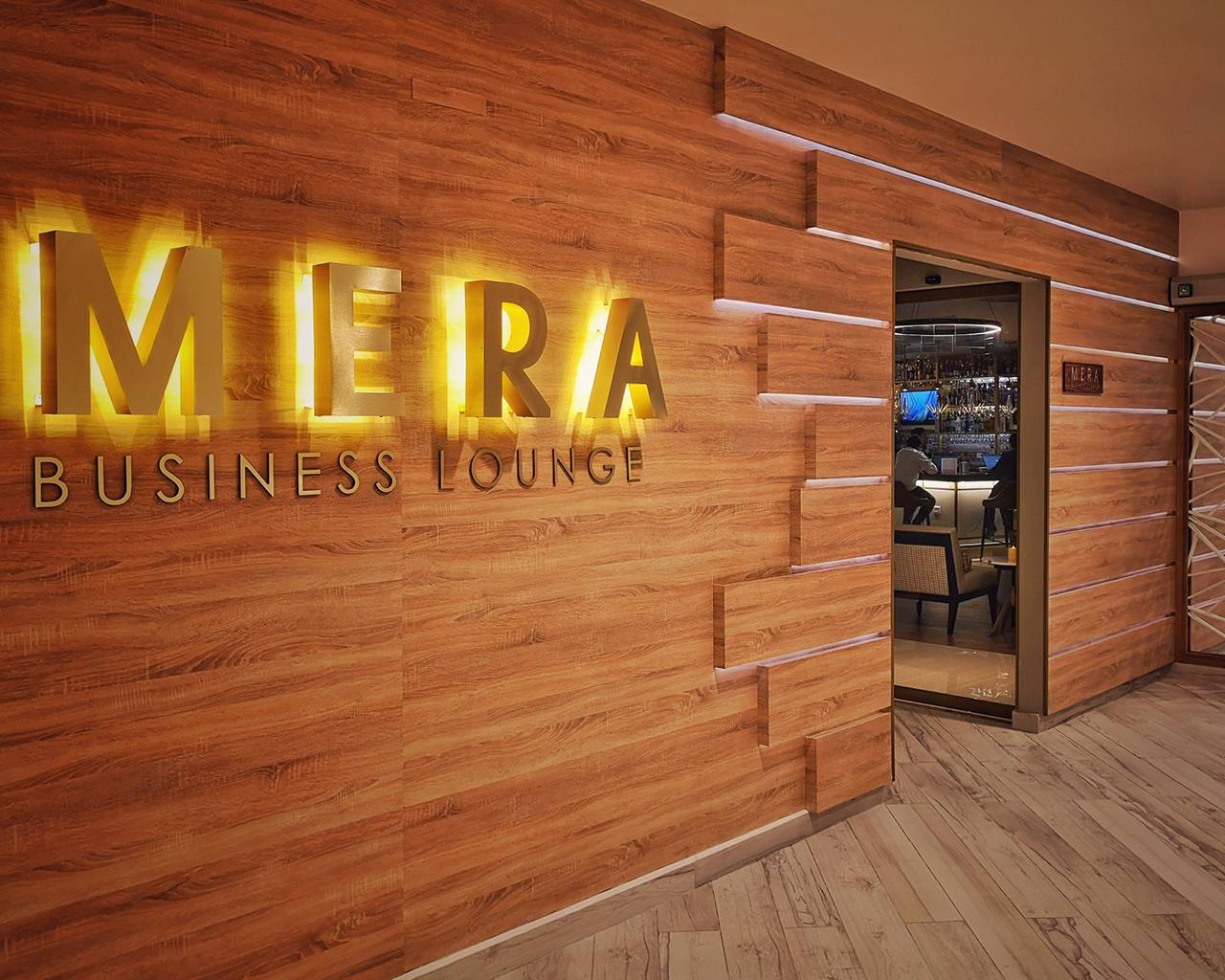 The Mera Business Lounge Cancun entrance with a wood-paneled feature wall and illuminated “MERA Business Lounge” signage. An open doorway reveals the bar area inside, with warm lighting and marble-style flooring continuing into the space. The Mera Business Lounge Cancun entrance with a wood-paneled feature wall and illuminated “MERA Business Lounge” signage. An open doorway reveals the bar area inside, with warm lighting and marble-style flooring continuing into the space.