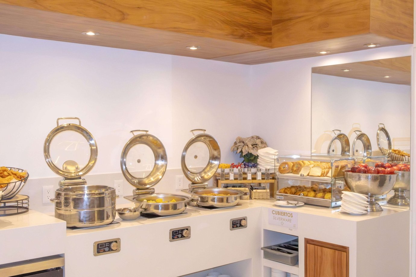 A buffet counter in the Mera Roof Lounge Cancun featuring three stainless steel hot food trays with lids, alongside a display of bagels, sliced bread, and pastries in a glass case. A buffet counter in the Mera Roof Lounge Cancun featuring three stainless steel hot food trays with lids, alongside a display of bagels, sliced bread, and pastries in a glass case.