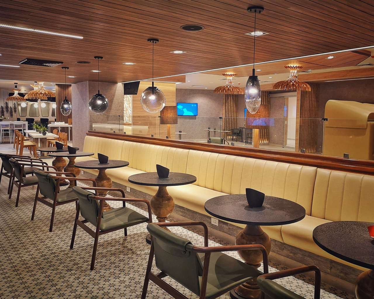 A seating area in the Mera Business Lounge Cancun with a long cream banquette, round dark tables, and wooden armchairs with green cushions. A seating area in the Mera Business Lounge Cancun with a long cream banquette, round dark tables, and wooden armchairs with green cushions.