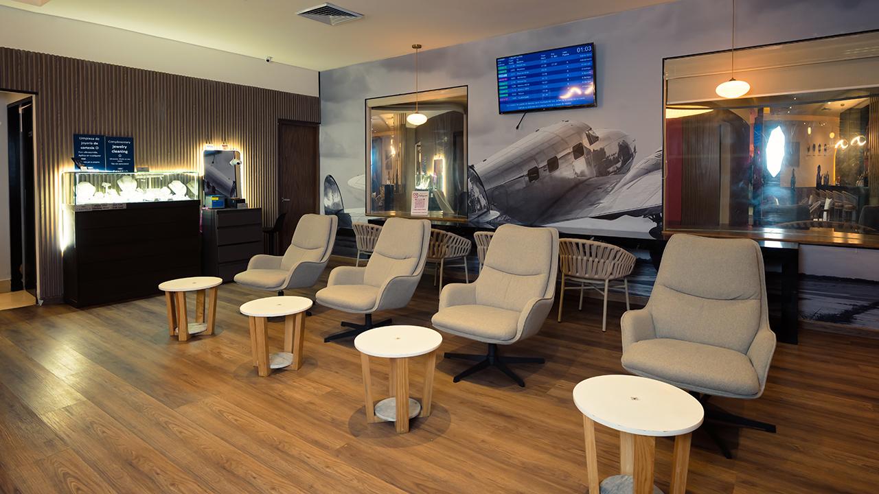 The Lounge T2 at Cancun International Airport seating area with upholstered armchairs, small round tables, wall art, and a flight monitor. The Lounge T2 at Cancun International Airport seating area with upholstered armchairs, small round tables, wall art, and a flight monitor.
