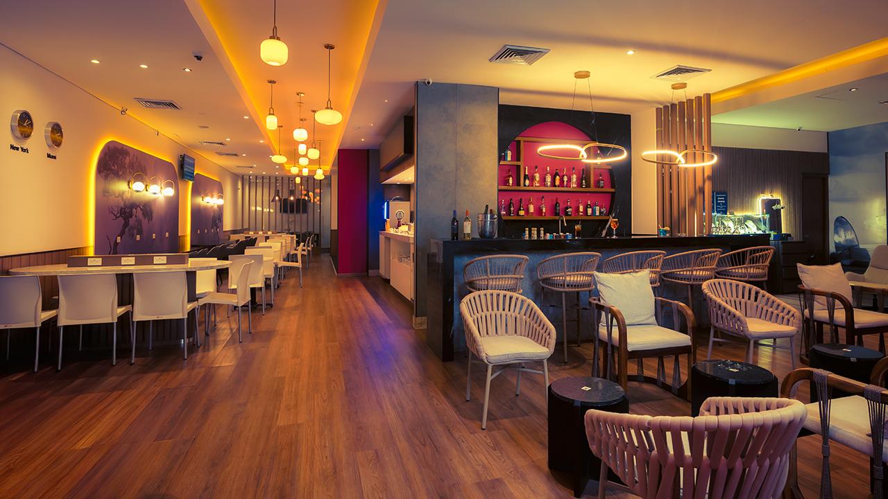 The Lounge T2 at Cancun International Airport bar and seating area with illuminated bar, high stools, dining tables, lounge chairs, and pendant lighting. The Lounge T2 at Cancun International Airport bar and seating area with illuminated bar, high stools, dining tables, lounge chairs, and pendant lighting.