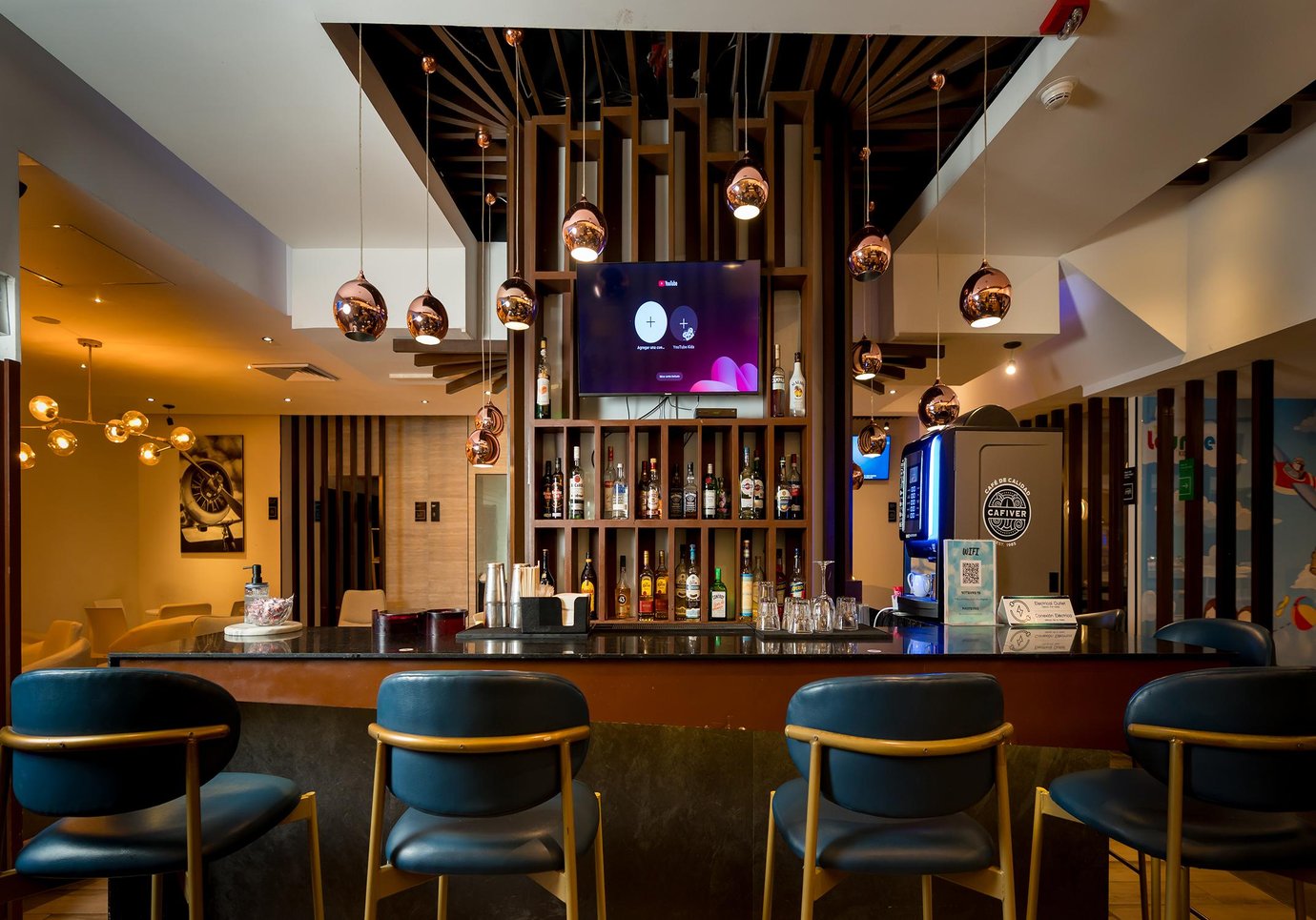 Mera Business Lounge Cancun bar area with a central counter, blue upholstered bar stools, and backlit shelving displaying liquor bottles. Features a wall-mounted TV above the bar, a coffee machine to the side, and multiple copper pendant lights hanging from a geometric wood ceiling structure. Mera Business Lounge Cancun bar area with a central counter, blue upholstered bar stools, and backlit shelving displaying liquor bottles. Features a wall-mounted TV above the bar, a coffee machine to the side, and multiple copper pendant lights hanging from a geometric wood ceiling structure.