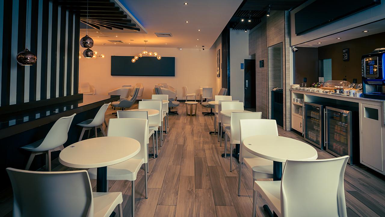 Mera Business Lounge Cancun seating and buffet area with rows of white round tables and matching chairs on wood flooring. Mera Business Lounge Cancun seating and buffet area with rows of white round tables and matching chairs on wood flooring.