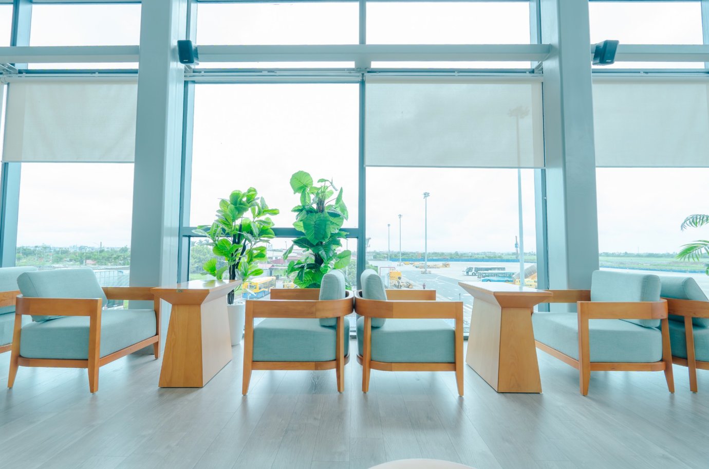 Row of light-blue cushioned armchairs with wooden frames and small side tables positioned beside large floor-to-ceiling windows with roller blinds, overlooking the airport apron at SH Premium Lounge, Cat Bi International Airport (HPH).