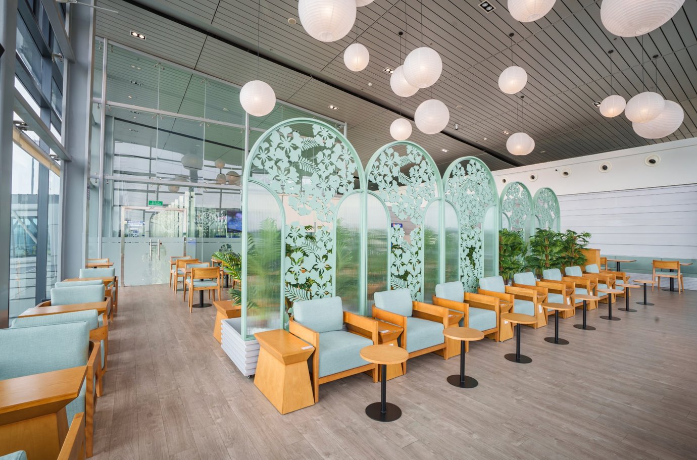 Seating area at SH Premium Lounge, Cat Bi International Airport (HPH), with rows of wooden armchairs, small round side tables, decorative frosted glass partitions with floral patterns, and hanging globe lights overhead.