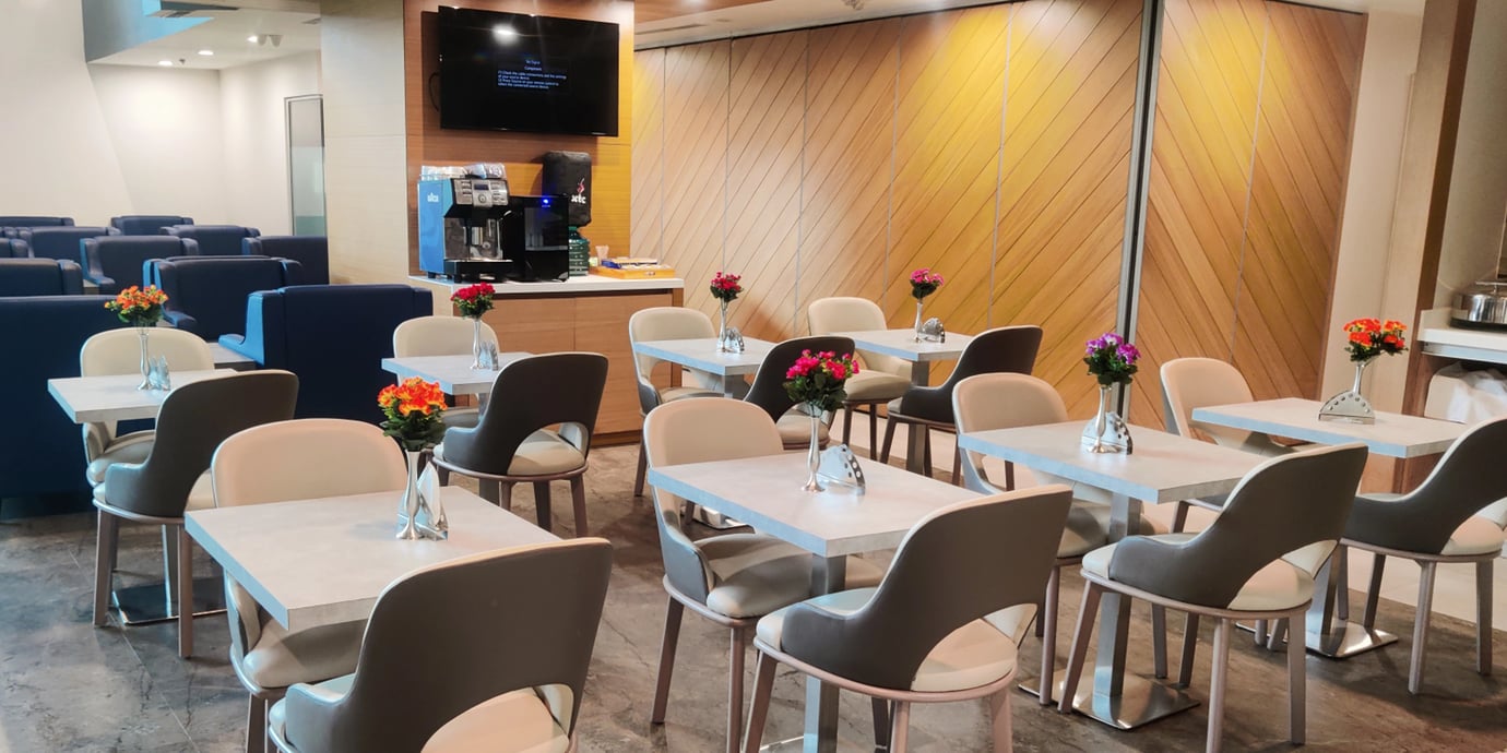 Dining area at Aviserv East Lounge Mumbai Airport featuring modern tables, cushioned chairs, floral décor, and a self-service coffee station. Dining area at Aviserv East Lounge Mumbai Airport featuring modern tables, cushioned chairs, floral décor, and a self-service coffee station.