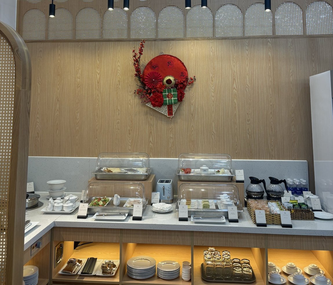 SH Premium Lounge buffet area at Con Dao Airport (VCS), featuring a compact yet well-presented spread of light hot dishes, salads, pastries, and beverages, set against warm wood finishes, woven panel accents, and soft lighting