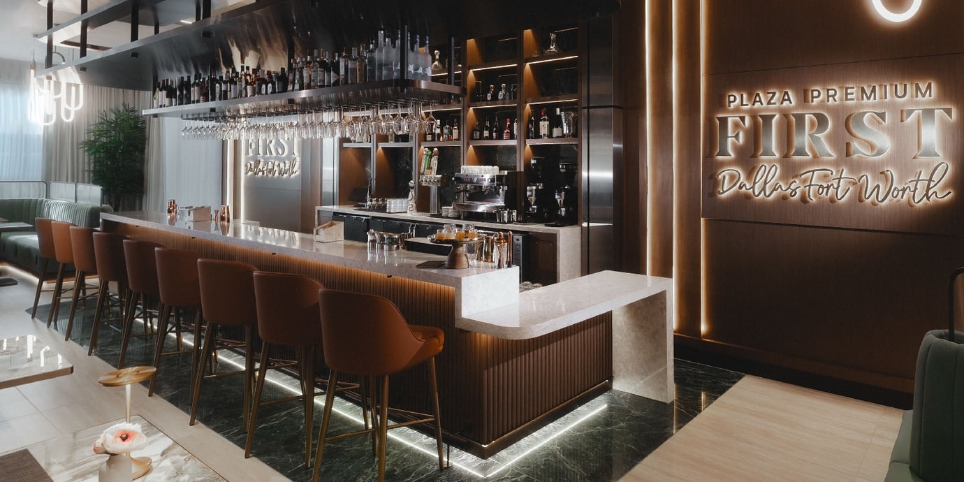 Bar area inside the Plaza Premium First Lounge at Dallas Fort Worth Airport. The design features a long marble-topped cocktail bar with warm underlighting, high-back leather bar stools, suspended glassware, and an extensive selection of premium spirits and wines. Bar area inside the Plaza Premium First Lounge at Dallas Fort Worth Airport. The design features a long marble-topped cocktail bar with warm underlighting, high-back leather bar stools, suspended glassware, and an extensive selection of premium spirits and wines.