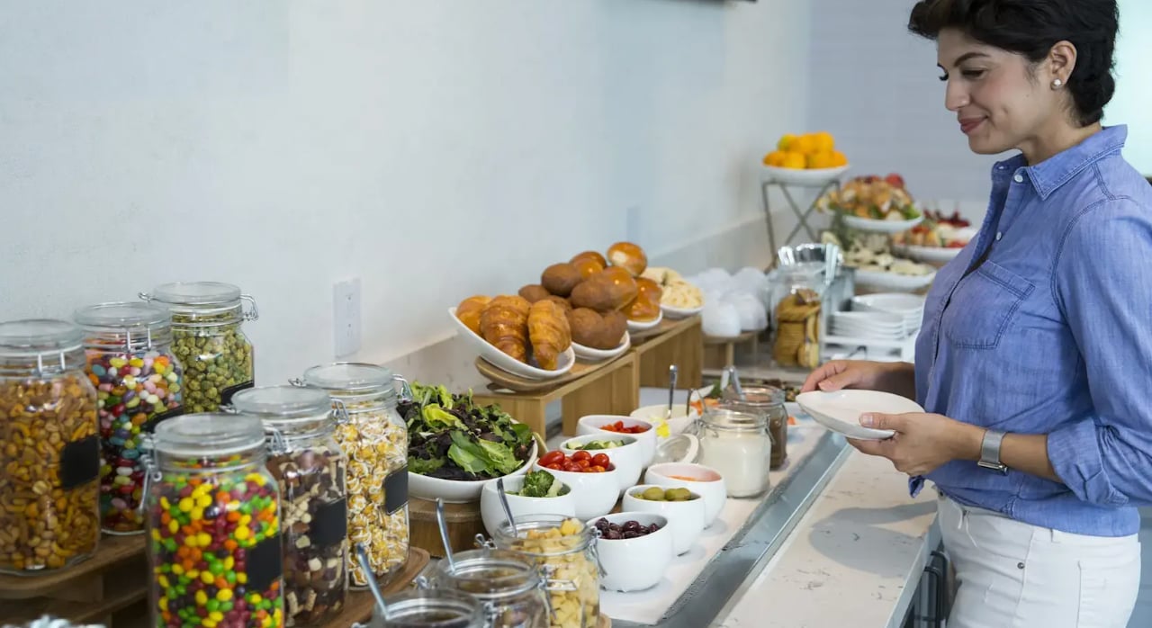 Traveller standing at a self-serve buffet inside The Club DFW airport lounge, selecting from a variety of snacks, pastries, salads, and toppings. Traveller standing at a self-serve buffet inside The Club DFW airport lounge, selecting from a variety of snacks, pastries, salads, and toppings.