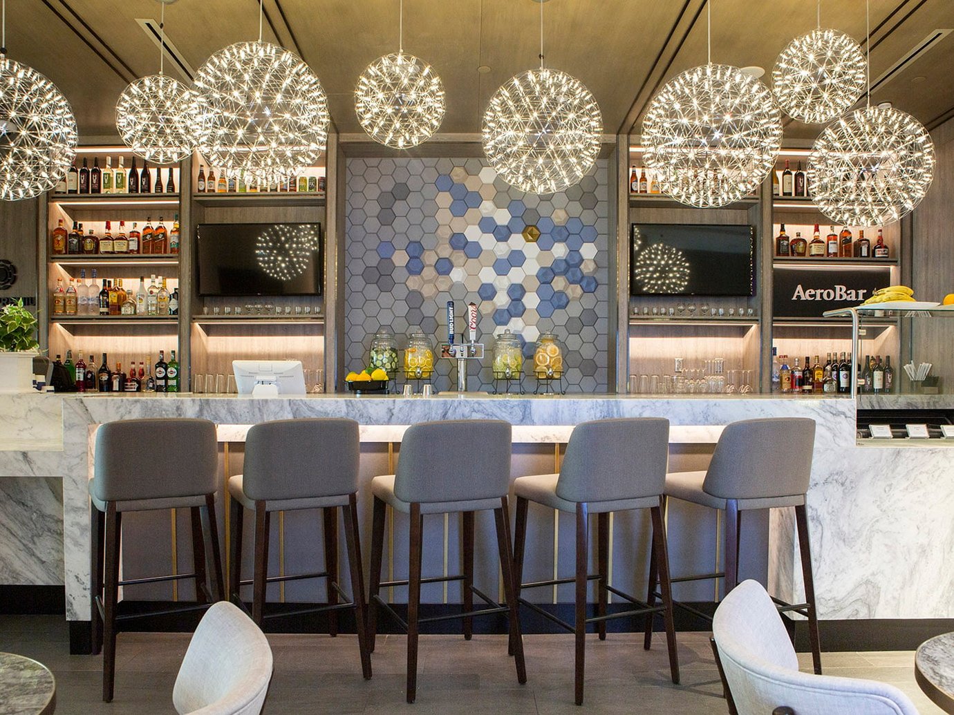 AeroBar inside the Plaza Premium Lounge Terminal E at DFW Airport. The bar features a polished white marble counter with high-back seating, illuminated liquor shelving, dual flat-screen displays, and geometric hexagonal wall tile accents. AeroBar inside the Plaza Premium Lounge Terminal E at DFW Airport. The bar features a polished white marble counter with high-back seating, illuminated liquor shelving, dual flat-screen displays, and geometric hexagonal wall tile accents.
