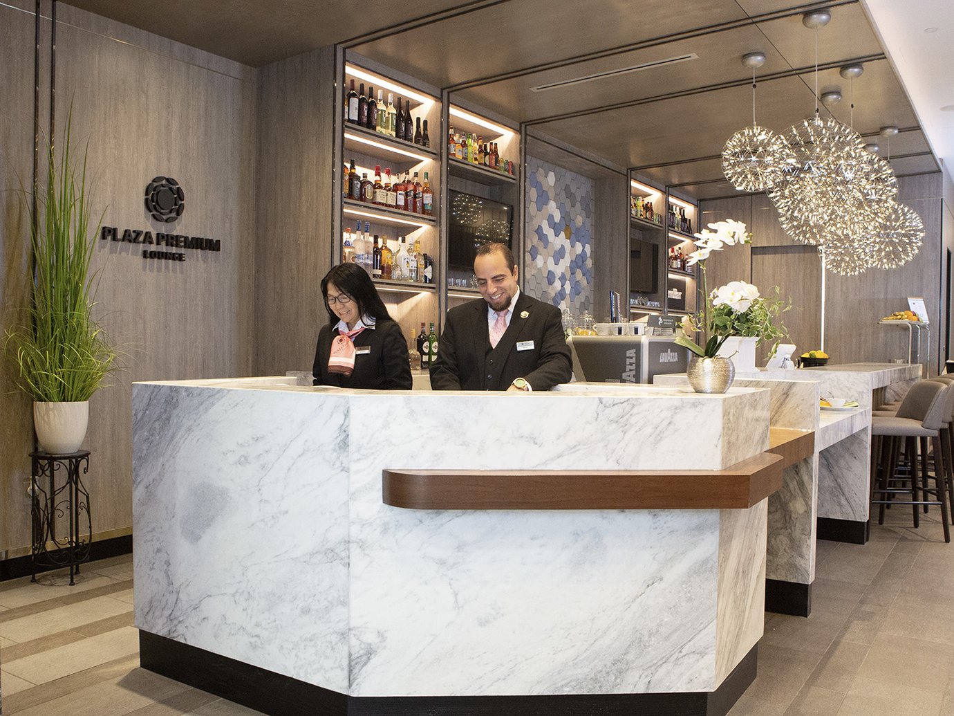 The reception and welcome area of the Plaza Premium Lounge in Terminal E at Dallas Fort Worth Airport. The lounge features a contemporary hospitality design with a curved white marble reception desk, warm wood paneling, illuminated liquor displays, and decorative pendant lighting The reception and welcome area of the Plaza Premium Lounge in Terminal E at Dallas Fort Worth Airport. The lounge features a contemporary hospitality design with a curved white marble reception desk, warm wood paneling, illuminated liquor displays, and decorative pendant lighting