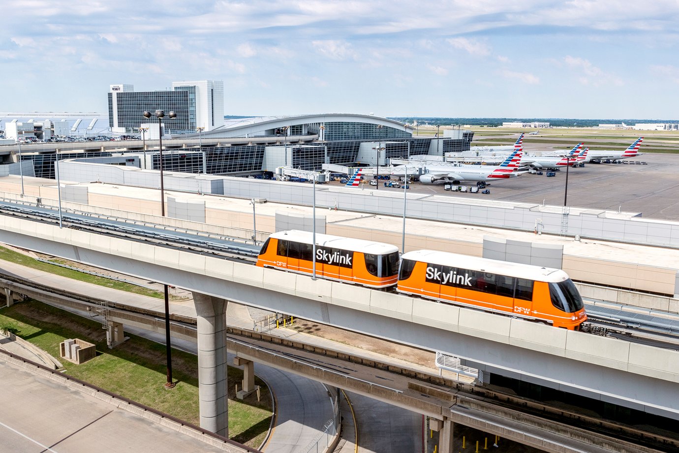 DFW Airport Skylink train connecting terminals at Dallas Fort Worth International Airport, with terminal buildings, aircraft gates, and airside operations visible in the background. DFW Airport Skylink train connecting terminals at Dallas Fort Worth International Airport, with terminal buildings, aircraft gates, and airside operations visible in the background.