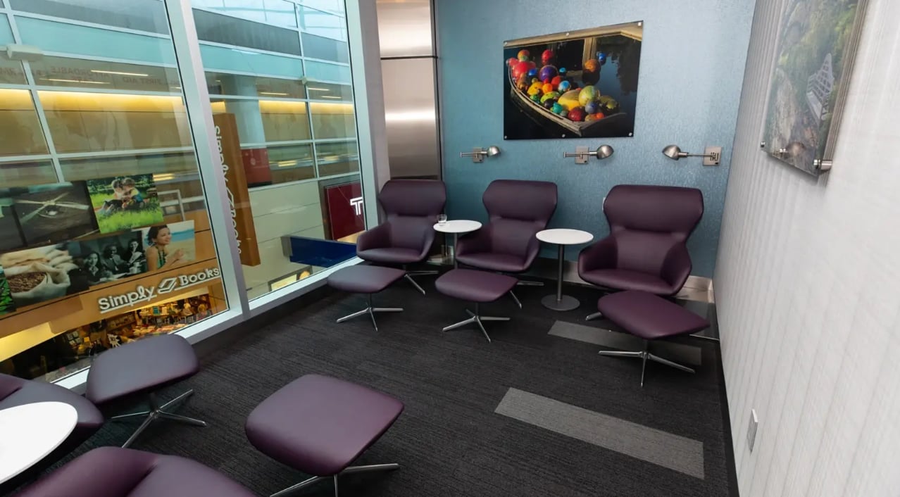 A quiet seating area inside The Club DFW airport lounge features modern purple lounge chairs with matching ottomans, small side tables, and individual reading lights. A quiet seating area inside The Club DFW airport lounge features modern purple lounge chairs with matching ottomans, small side tables, and individual reading lights.