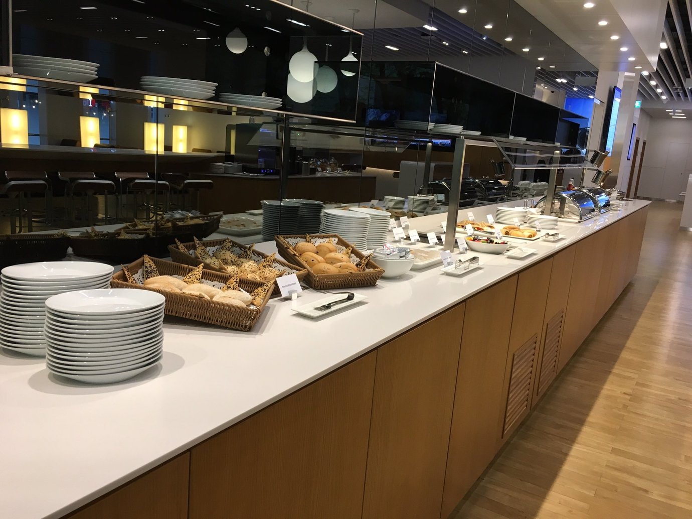 Buffet counter inside the Lufthansa Senator Lounge at Dubai International Airport Terminal 1, displaying a spread of breads, pastries, hot dishes, and plates neatly stacked on a long white counter with a sleek modern design and soft lighting. Buffet counter inside the Lufthansa Senator Lounge at Dubai International Airport Terminal 1, displaying a spread of breads, pastries, hot dishes, and plates neatly stacked on a long white counter with a sleek modern design and soft lighting.