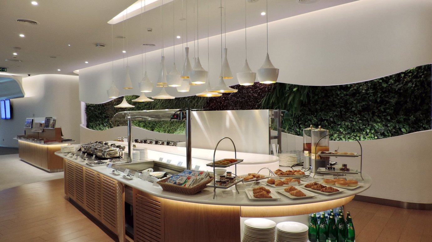 Buffet area inside the SkyTeam Lounge at Dubai International Airport Terminal 1, featuring a modern counter with pastries, salads, and hot dishes under elegant pendant lighting and a lush green wall backdrop. Buffet area inside the SkyTeam Lounge at Dubai International Airport Terminal 1, featuring a modern counter with pastries, salads, and hot dishes under elegant pendant lighting and a lush green wall backdrop.