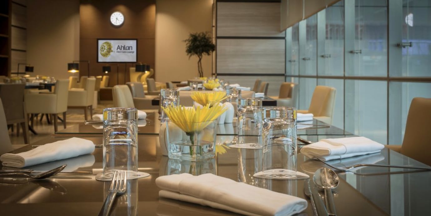 Elegant dining setup inside the Ahlan First Class Lounge at Dubai International Airport Terminal 1, featuring neatly arranged tables with glassware, white linens, and fresh yellow flowers in a refined, tranquil atmosphere. Elegant dining setup inside the Ahlan First Class Lounge at Dubai International Airport Terminal 1, featuring neatly arranged tables with glassware, white linens, and fresh yellow flowers in a refined, tranquil atmosphere.