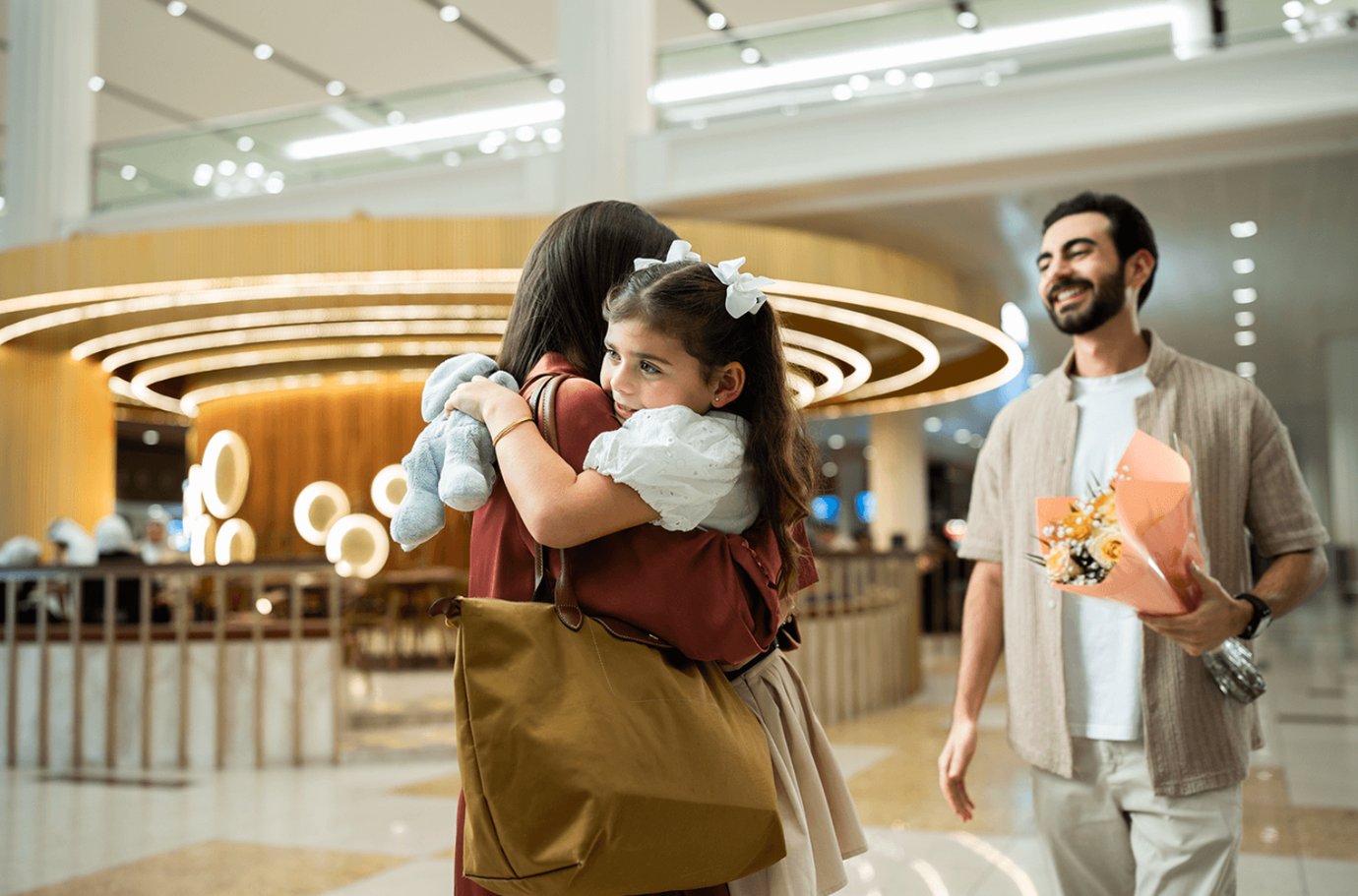 A young girl hugs a woman tightly at Dubai International Airport Terminal 1 arrivals, holding a stuffed toy while a man approaches smiling with a bouquet of flowers. A young girl hugs a woman tightly at Dubai International Airport Terminal 1 arrivals, holding a stuffed toy while a man approaches smiling with a bouquet of flowers.