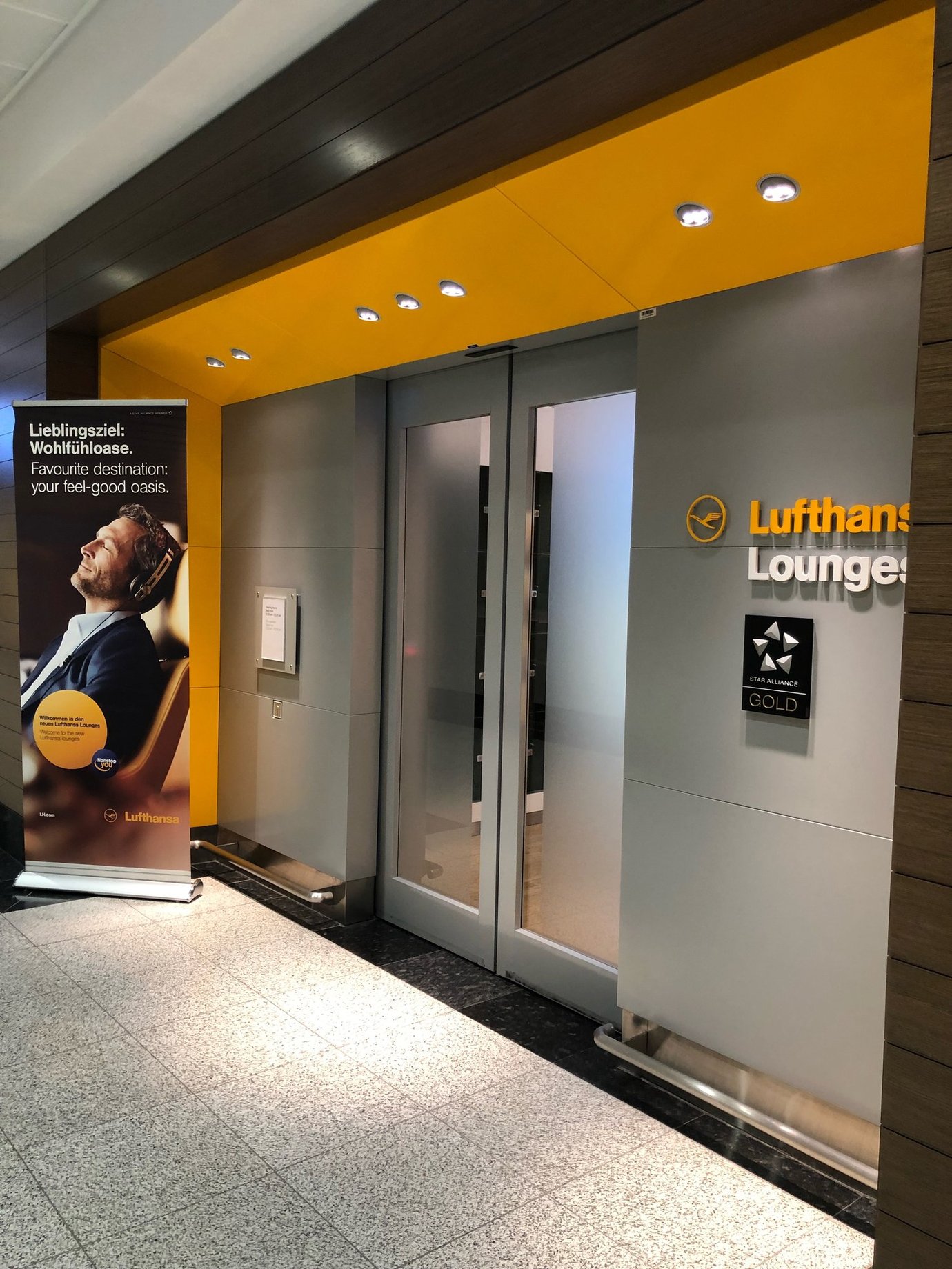 Entrance to the Lufthansa Business Lounge at Dubai International Airport Terminal 1, featuring the airline’s signature yellow branding, frosted glass doors, and Star Alliance Gold signage beside a promotional display. Entrance to the Lufthansa Business Lounge at Dubai International Airport Terminal 1, featuring the airline’s signature yellow branding, frosted glass doors, and Star Alliance Gold signage beside a promotional display.