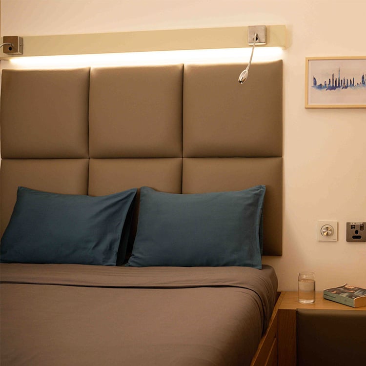 Sleepover pod room at Dubai International Airport featuring a compact modern bed with a padded beige headboard, blue pillows, and a bedside table holding a glass of water and a book. Sleepover pod room at Dubai International Airport featuring a compact modern bed with a padded beige headboard, blue pillows, and a bedside table holding a glass of water and a book.