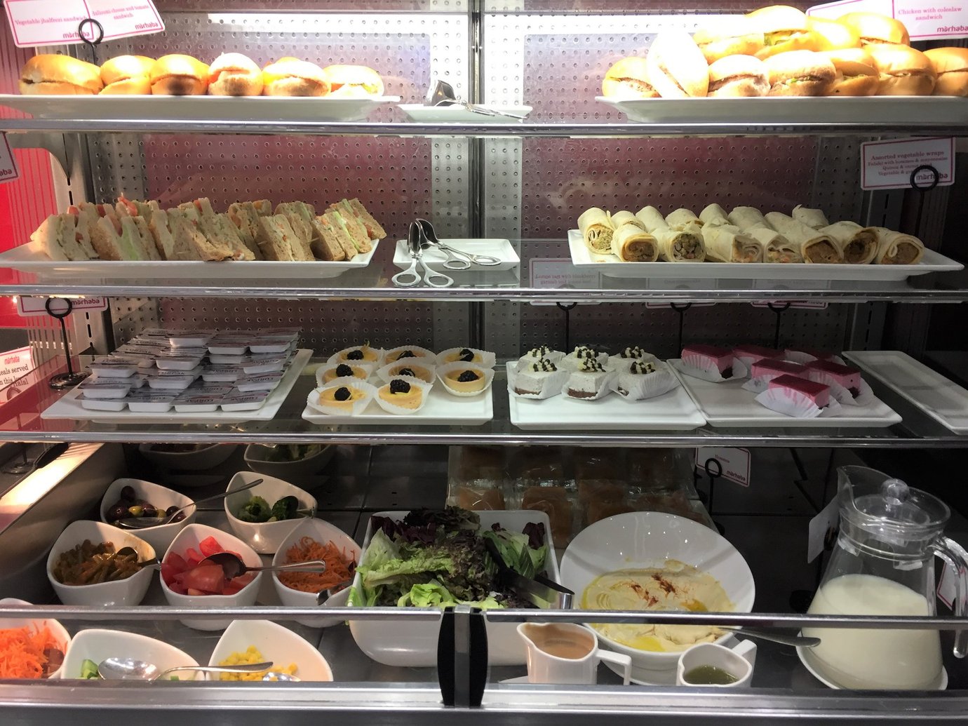 Buffet display at Marhaba Lounge in Dubai Airport Terminal 2 showing assorted sandwiches, wraps, pastries, salads, and desserts in a well-lit self-service dining area. Buffet display at Marhaba Lounge in Dubai Airport Terminal 2 showing assorted sandwiches, wraps, pastries, salads, and desserts in a well-lit self-service dining area.