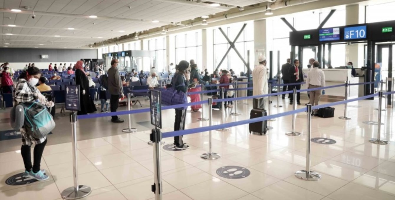 Passengers queueing at Dubai Airport Terminal 2 Gate F10, maintaining distance in a bright departure area with seating, information screens, and boarding gates visible. Passengers queueing at Dubai Airport Terminal 2 Gate F10, maintaining distance in a bright departure area with seating, information screens, and boarding gates visible.