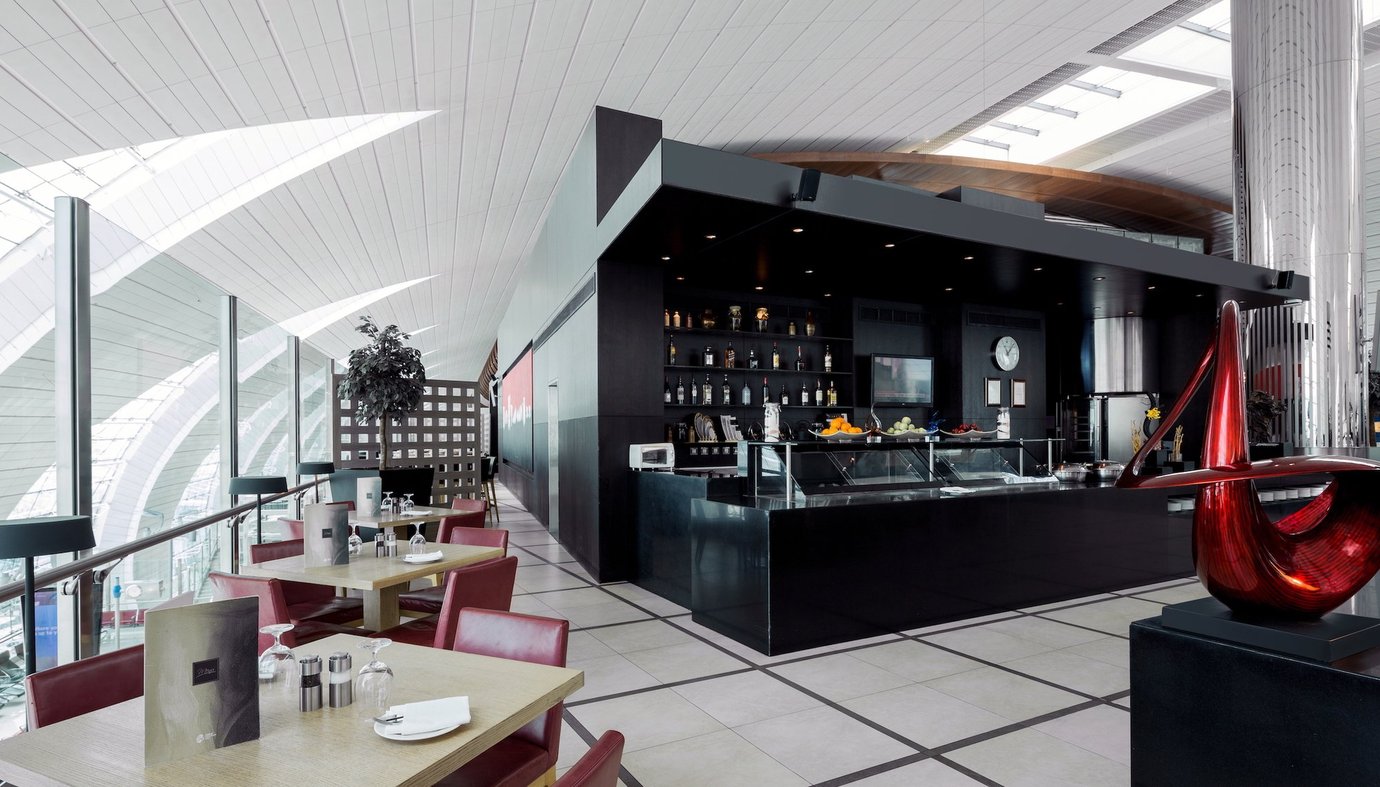 Buffet and bar area inside the Ahlan Lounge at Dubai International Airport Terminal 3 Concourse C, featuring sleek black interiors, red seating, and large windows overlooking the terminal for a modern upscale ambiance.