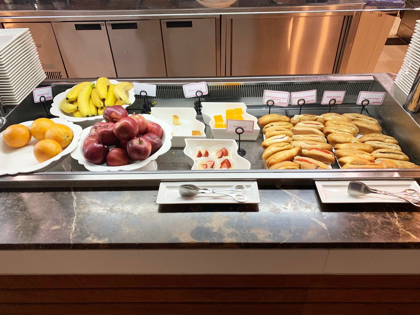 Buffet counter inside the Marhaba Lounge at Dubai International Airport Terminal 3 Concourse B, featuring a selection of fresh fruit, cheese cubes, and assorted sandwiches arranged neatly on a marble countertop.