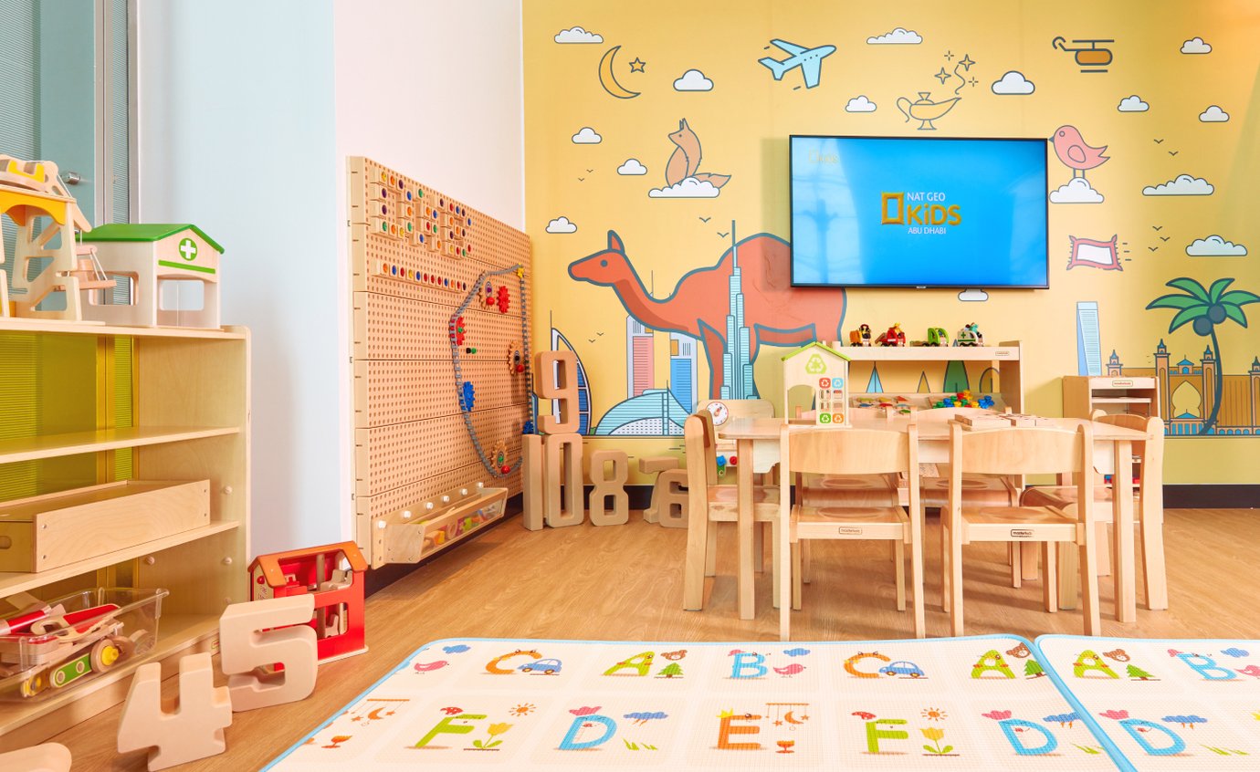 Children’s play area inside the Plaza Premium Lounge at Dubai International Airport Terminal 3, featuring colorful wall art, educational toys, wooden play furniture, and a Nat Geo Kids screen for entertainment.