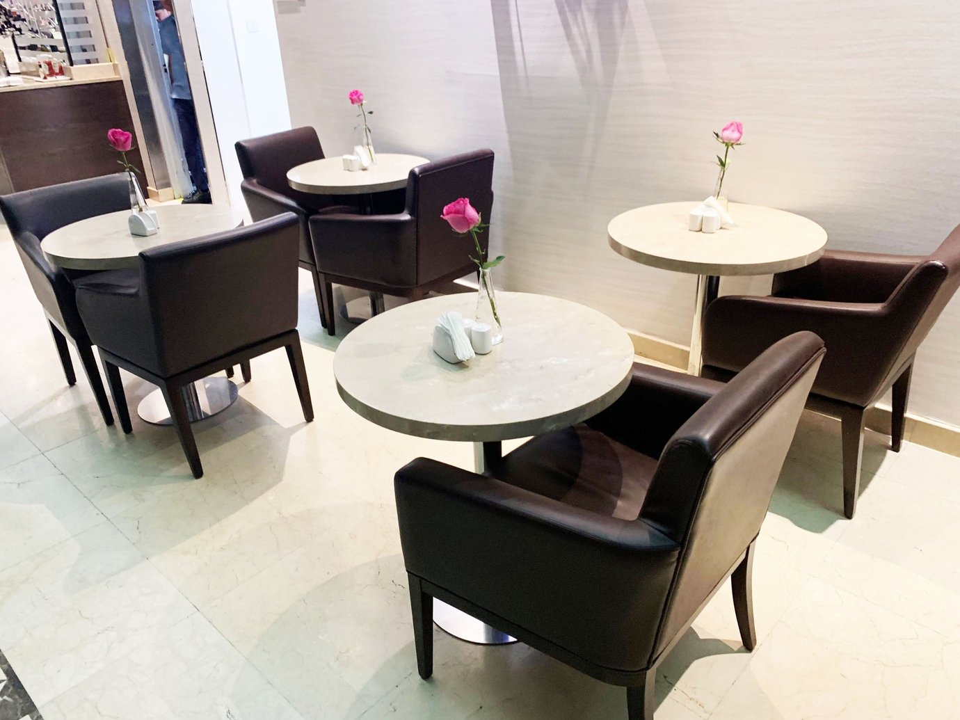 Dining area inside the Marhaba Lounge at Dubai International Airport Terminal 3 Concourse C, featuring dark leather chairs, round marble tables, and pink rose vases creating a cozy, elegant atmosphere