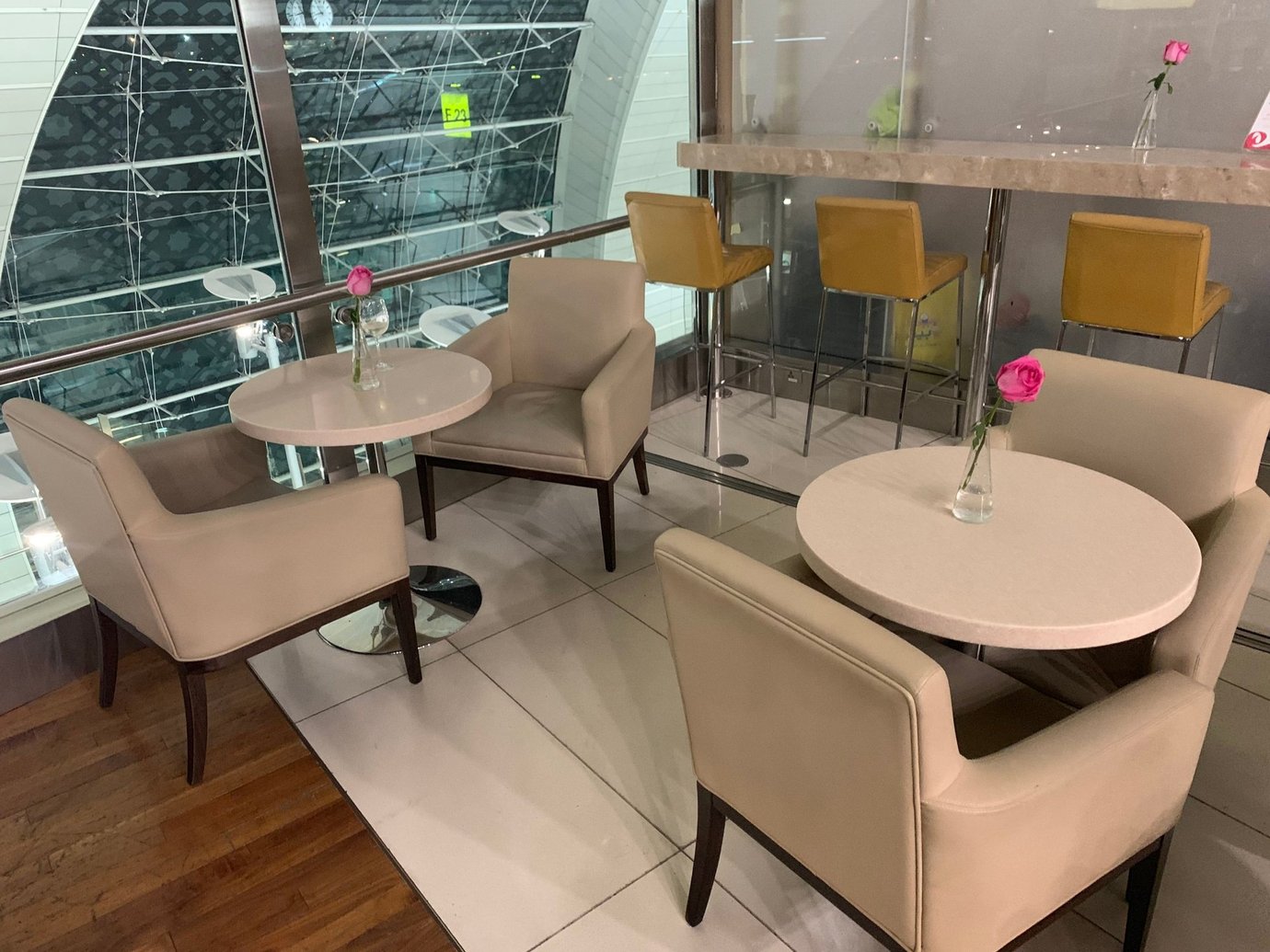 Dining seating area inside the Marhaba Lounge at Dubai International Airport Terminal 3 Concourse B, featuring beige leather chairs, round marble tables with single rose vases, and elevated bar seating overlooking the terminal’s glass architecture.