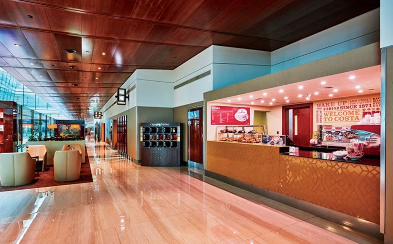Costa Coffee counter inside the Emirates Business Class Lounge at Concourse B, Dubai International Airport Terminal 3, featuring a warm wood-paneled ceiling, polished marble floors, beige leather seating, and soft ambient lighting.