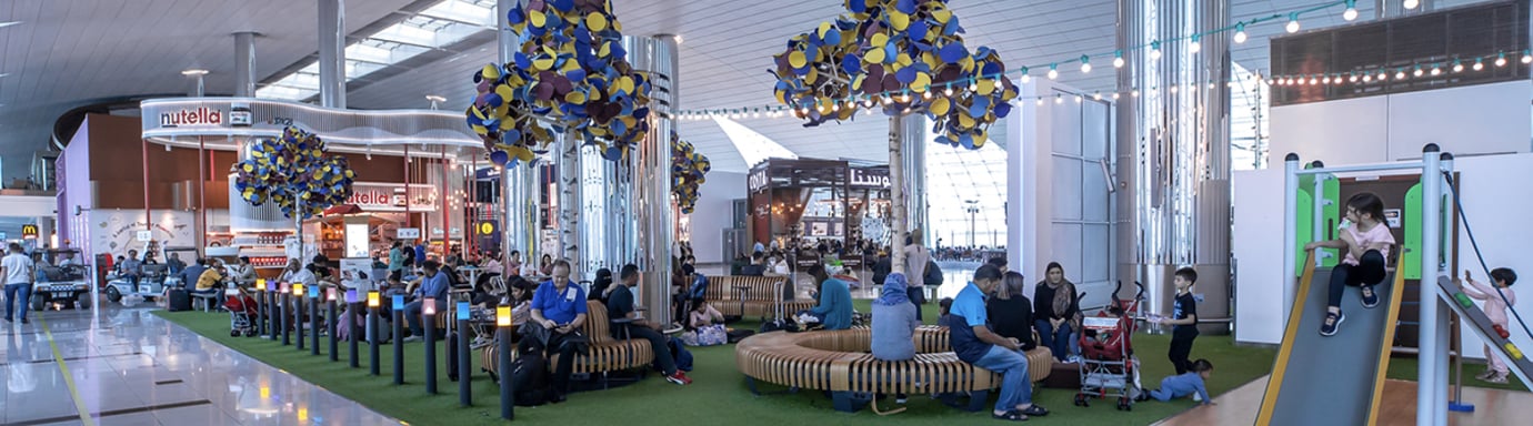Family Zone at Dubai International Airport’s Terminal 3, featuring a spacious seating area with modern wooden benches and colorful tree-like sculptures. Families relax while children play on a small slide and play area to the right.