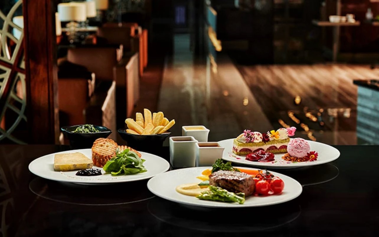 Close-up of elegant à la carte dishes served at the Emirates First Class Lounge in Dubai International Airport’s Terminal 3. Plates feature premium entrées such as grilled steak with asparagus and roasted tomatoes, puff pastry with greens, and an artfully plated dessert.