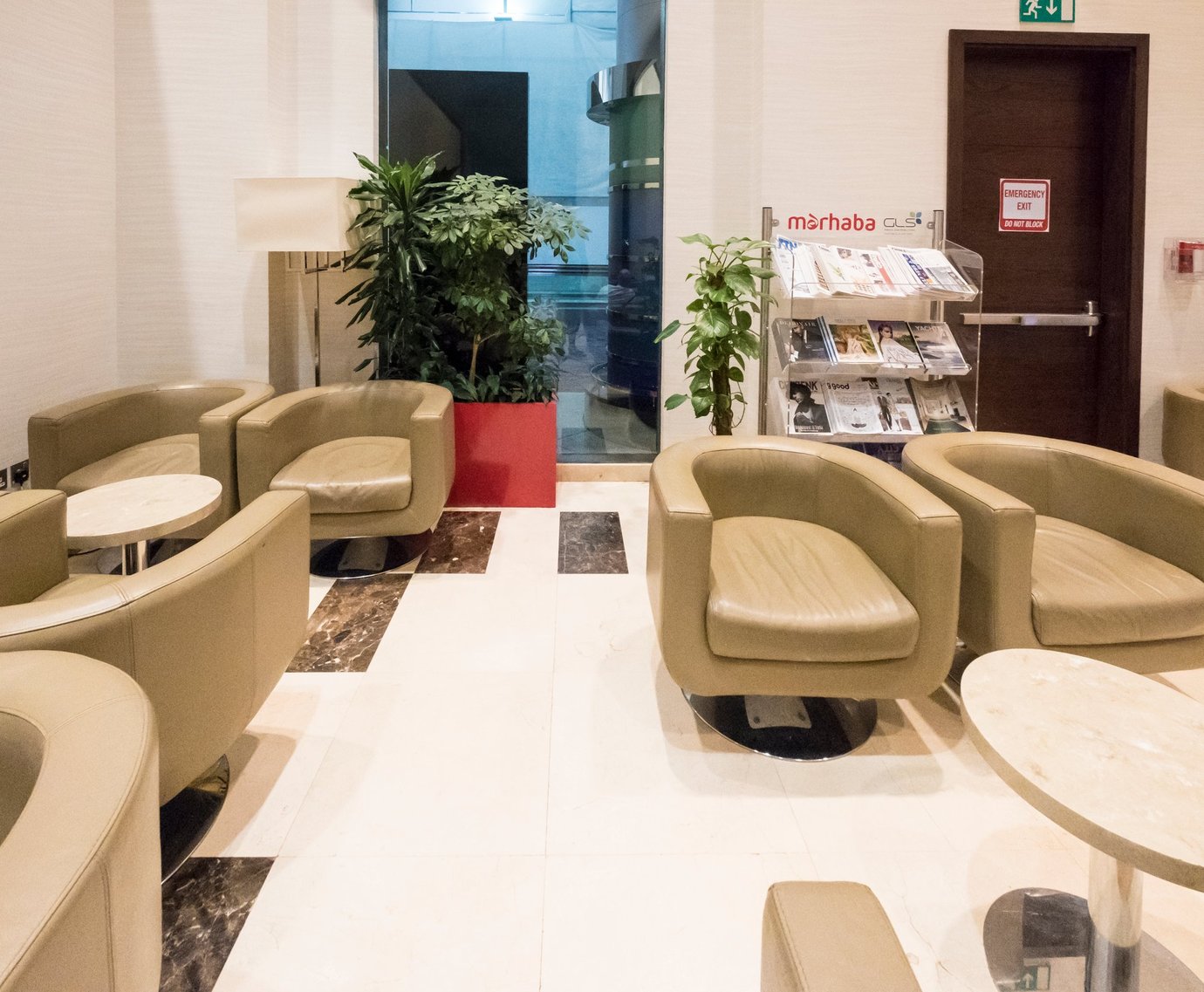 Seating area inside the Marhaba Lounge at Dubai International Airport Terminal 3 Concourse C, featuring beige leather armchairs, marble tables, indoor plants, and a magazine stand offering reading materials for guests.