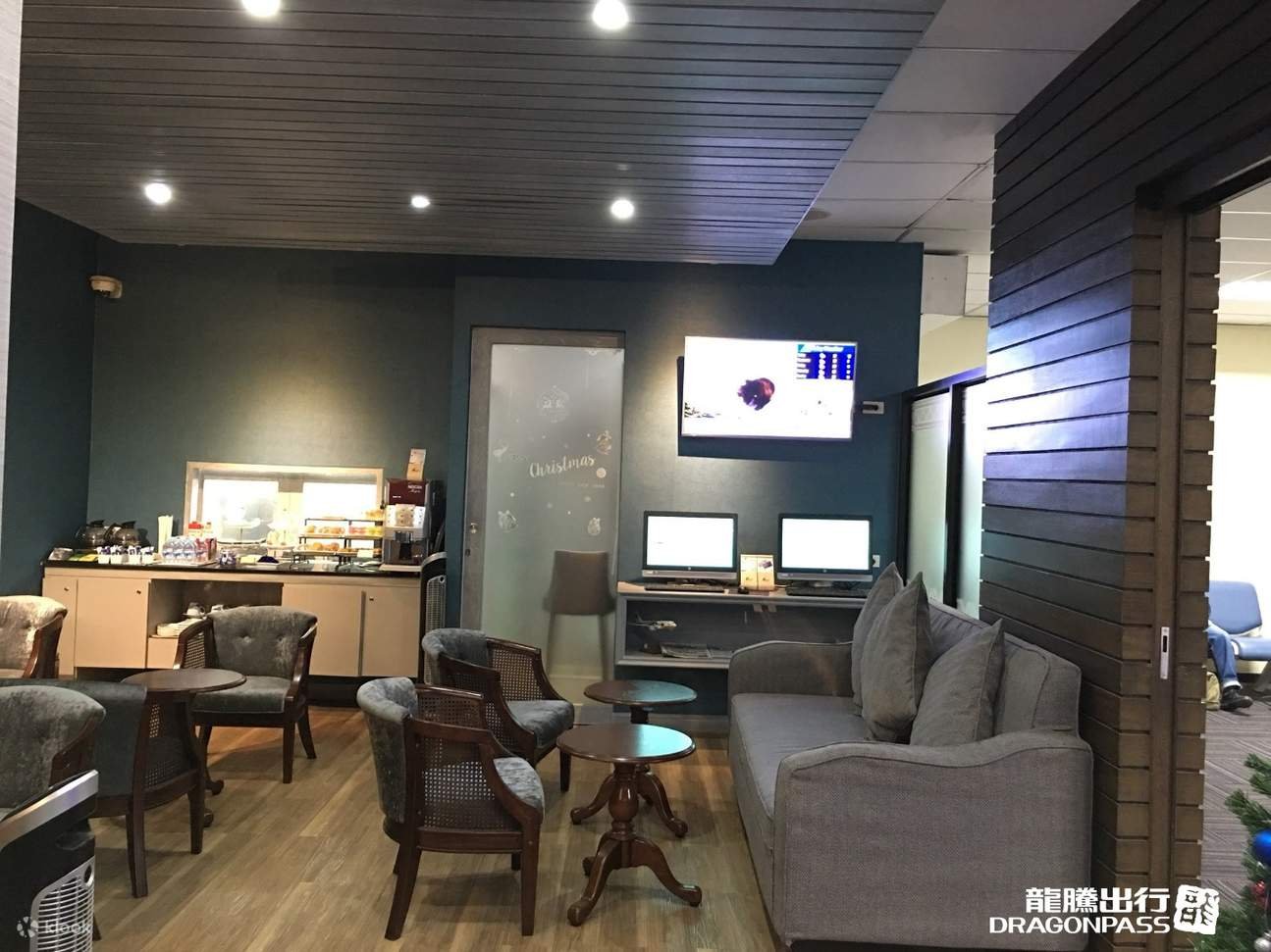 Chiang Rai Airport lounge seating area featuring cushioned armchairs, a grey sofa, coffee tables, refreshments counter with snacks and drinks, computer workstations, and a TV displaying flight information.