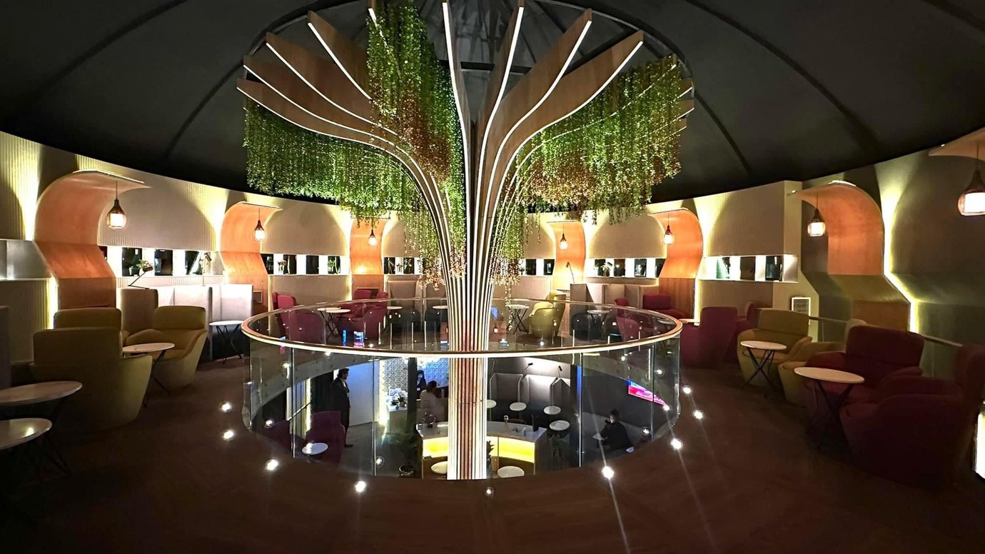Panoramic interior of Sky Majestic Lounge at Jinnah International Airport (KHI), showcasing circular mezzanine layout, central tree-inspired design with hanging greenery, and private seating pods
