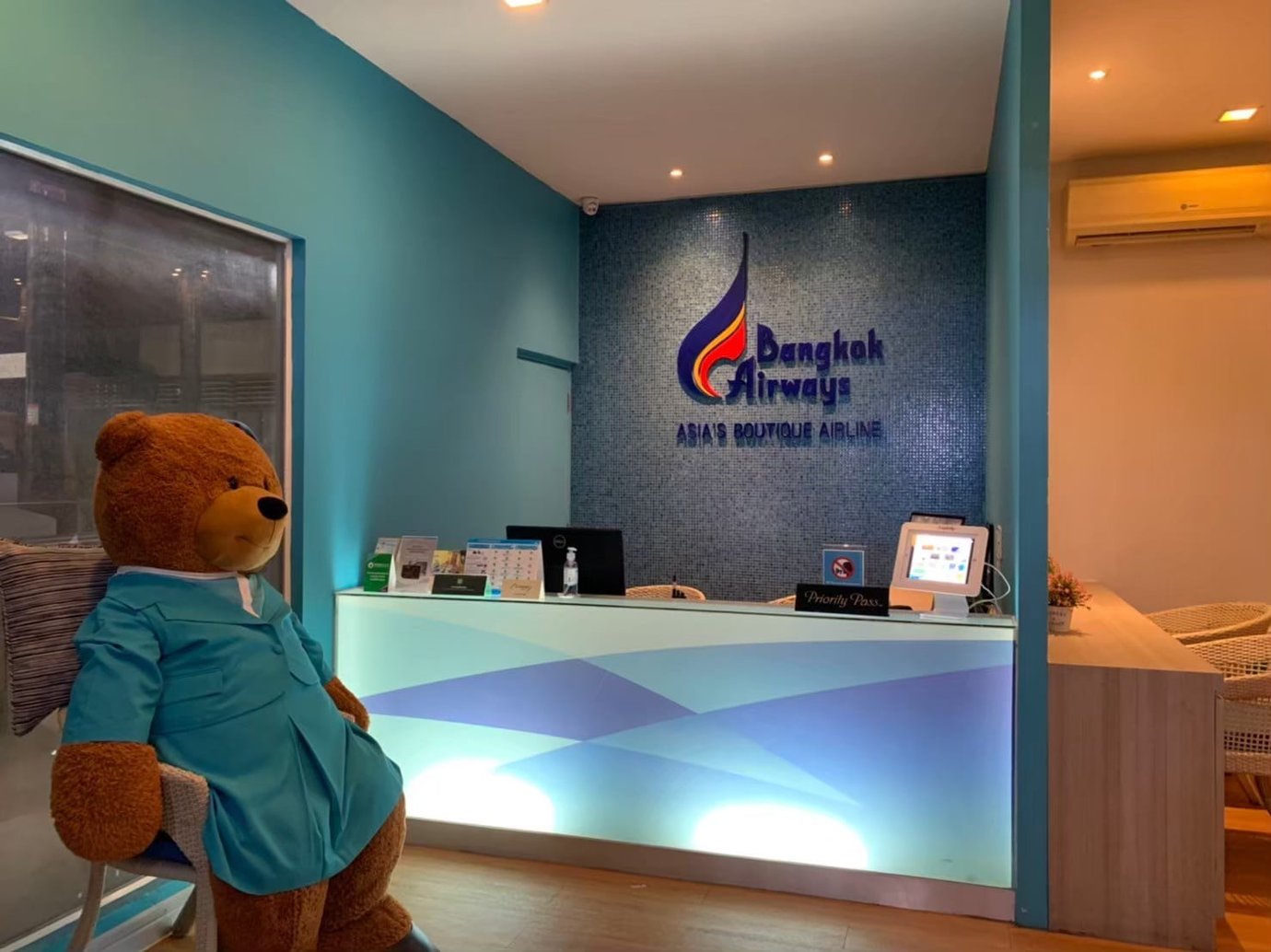 Reception desk at USM Blue Ribbon Lounge (International), featuring Bangkok Airways branding on a mosaic wall, a check-in counter with a tablet device, soft blue lighting, and a teddy bear statue dressed in a blue uniform beside the entrance.
