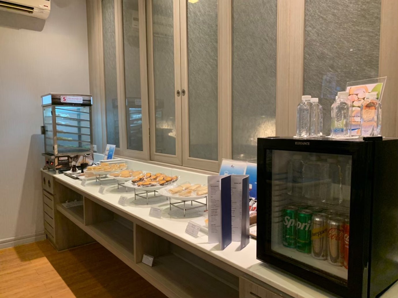 Buffet counter inside USM Blue Ribbon Lounge (International), displaying pastries and snacks on tiered trays, a food warmer unit, bottled water on top of a small refrigerated drinks fridge, and a menu stand on a long white countertop.