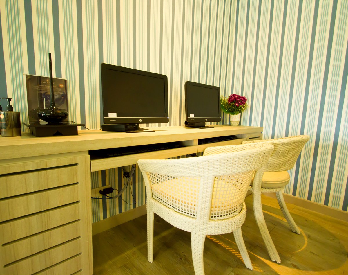 Computer workstation area inside USM Blue Ribbon Lounge (International), featuring two desktop monitors on a wooden desk, wicker chairs, and striped wall décor in a quiet corner workspace.