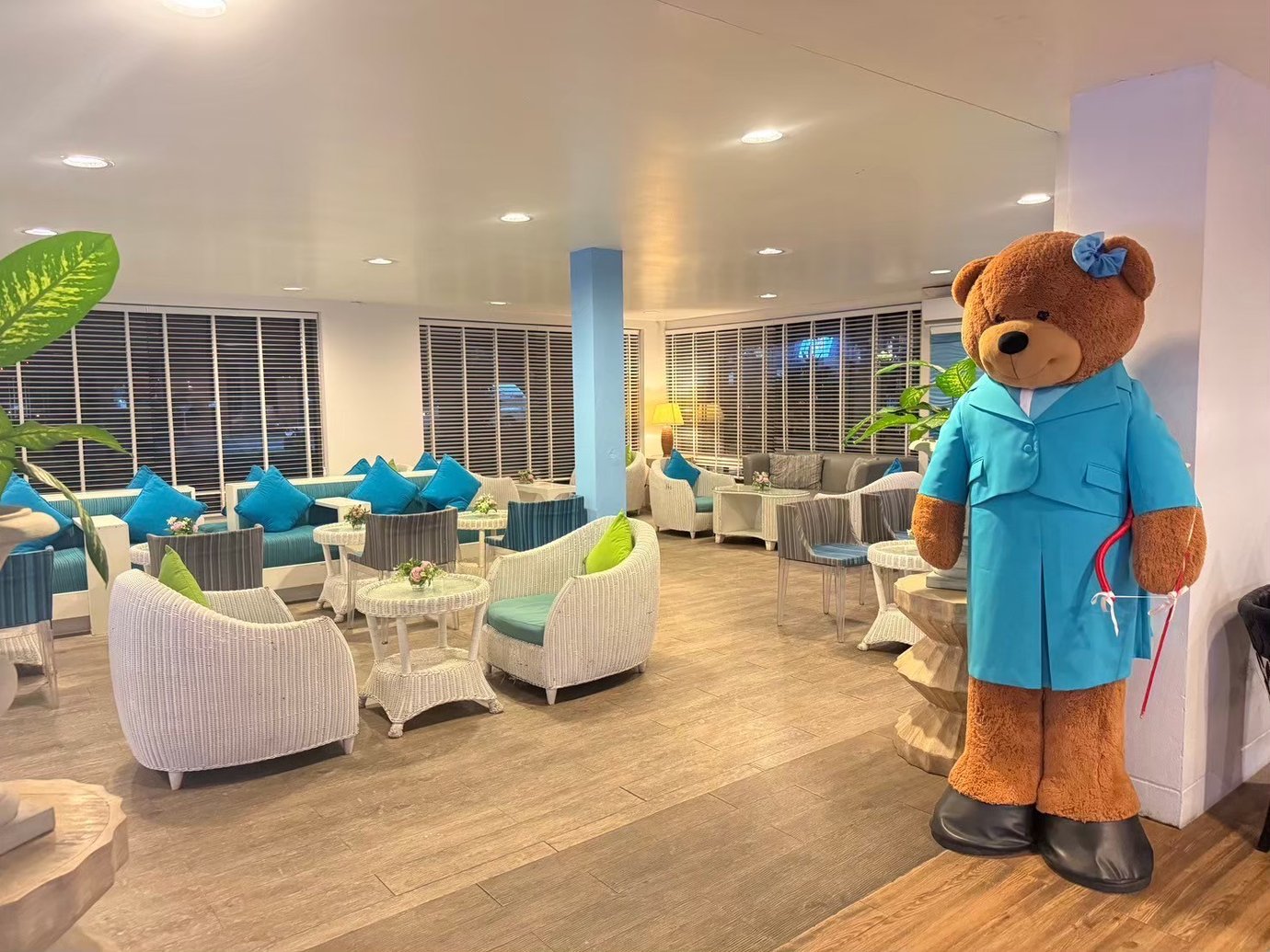 Seating area inside USM Blue Ribbon Lounge (Domestic), featuring white wicker chairs, round tables with blue and green cushions, and the Bangkok Airways signature bear next to a wall.