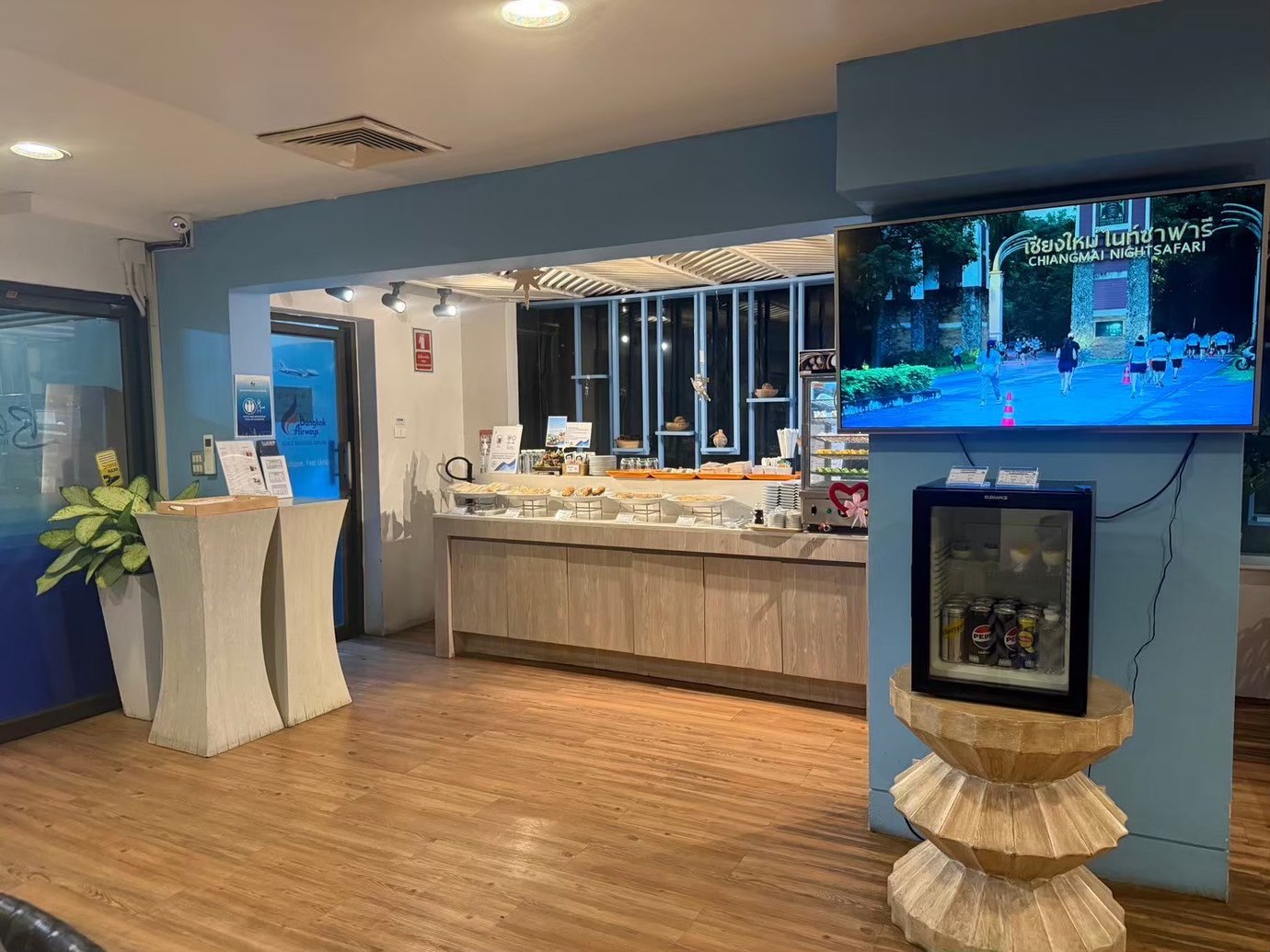 Buffet area inside USM Blue Ribbon Lounge (Domestic), featuring a self-service food counter with covered dishes, plates and drinks, a wall-mounted TV screen, a small refrigerated drinks unit, wooden flooring, and informational stands near the entrance.