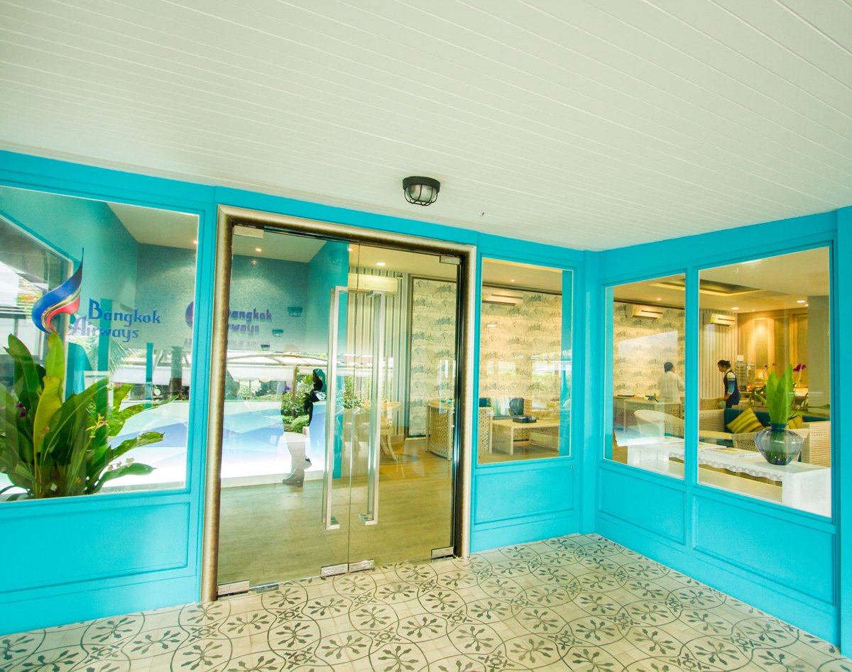 Entrance to USM Blue Ribbon Lounge (International), featuring glass double doors with Bangkok Airways branding, turquoise walls, patterned tile flooring, and a view into the lounge interior with seating and buffet area visible through the windows.