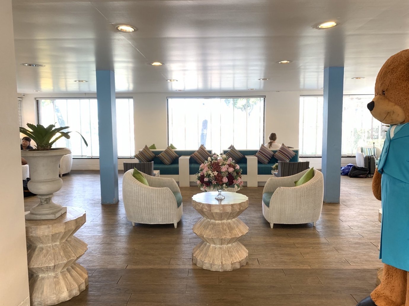 Central seating area inside USM Blue Ribbon Lounge (Domestic), featuring wicker armchairs around a decorative table with flowers, a sofa with patterned cushions along large windows with blinds, blue support columns, and a teddy bear statue positioned at the side.