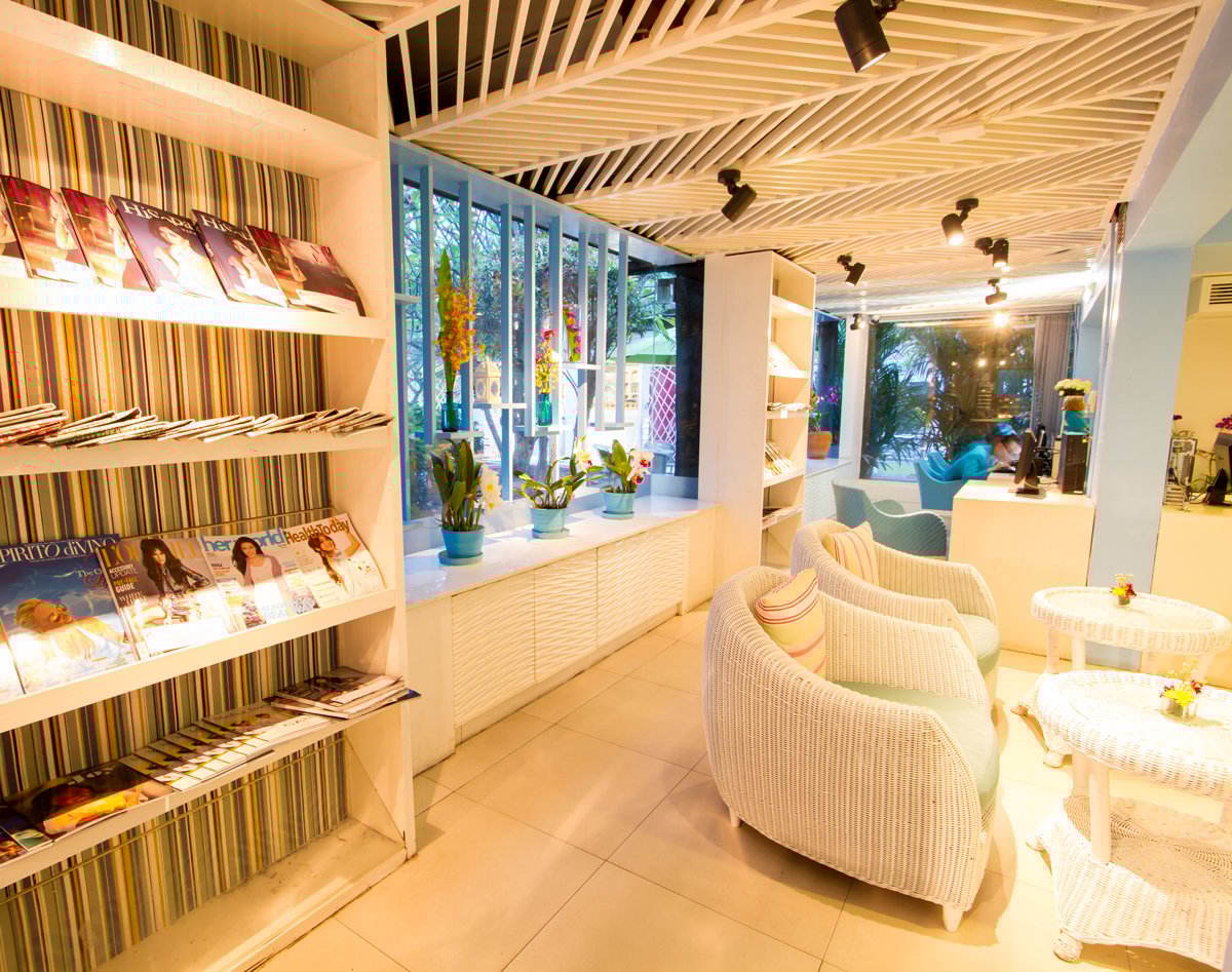 Reading and seating area inside USM Blue Ribbon Lounge (Domestic), with magazine display shelves, white wicker chairs and tables, potted plants along a window ledge, and a workspace with computer terminals in the background.