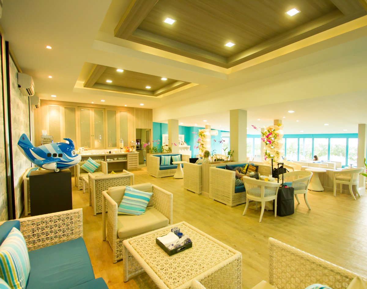 Main seating area inside USM Blue Ribbon Lounge (International), featuring wicker sofas and chairs with blue cushions, small tables, a buffet counter at the back, decorative plants, a model airplane display, and large windows letting in natural light.