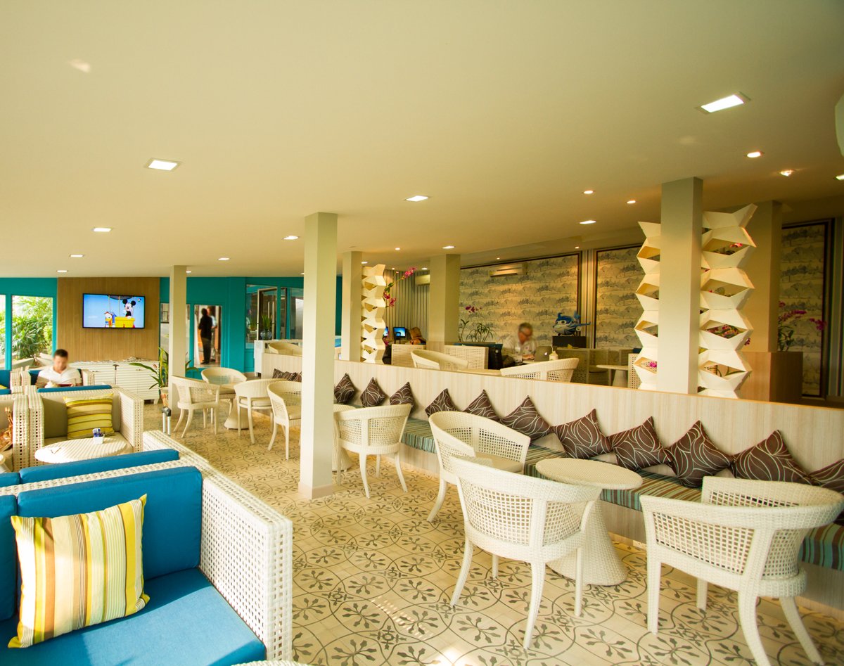 Seating layout inside USM Blue Ribbon Lounge (International), featuring white wicker chairs and tables, patterned tile flooring, bench seating with cushions along a partition wall, support columns, and a TV mounted on the wall in a bright, open lounge space.