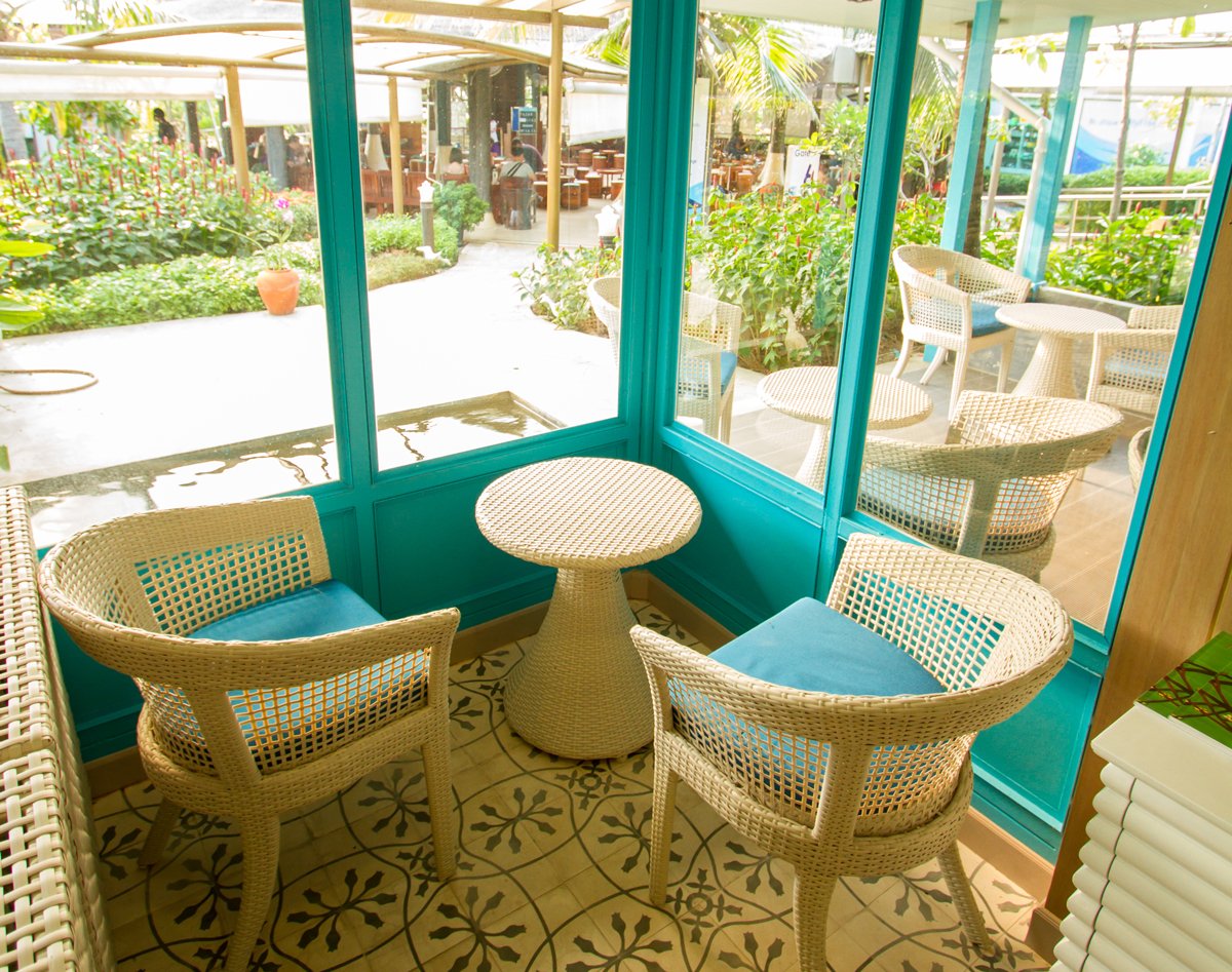 Window-side seating area in USM Blue Ribbon Lounge (International), featuring wicker chairs and a small round table inside a glass corner with views of a lush outdoor garden and shaded patio seating.