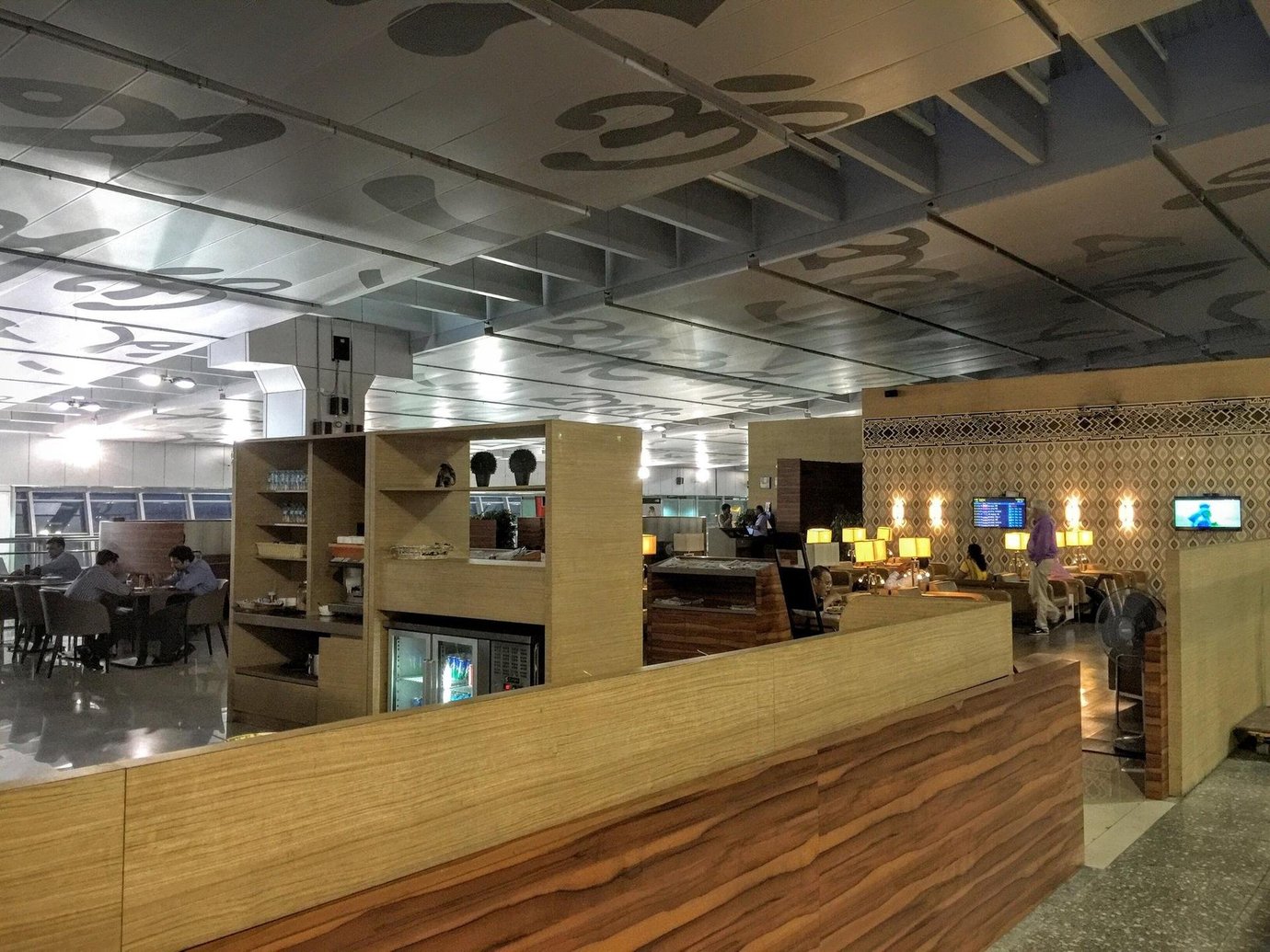 Travel Club Lounge at Kolkata Airport, featuring wooden accented partitions separating a dining area from a utensils and beverage station. A flight monitor is on the far wall, and warm yellow lamp lights give the space an inviting ambiance.